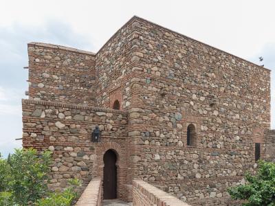 A low-angle view of a historic stone and brick fortress wall featuring an arched wooden door, small windows, and a mounted lantern, with green foliage in the foreground.