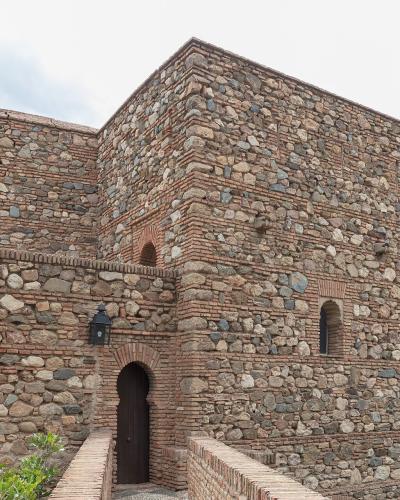 A low-angle view of a historic stone and brick fortress wall featuring an arched wooden door, small windows, and a mounted lantern, with green foliage in the foreground.