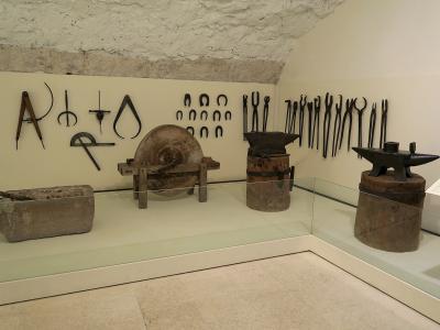 A museum display case containing antique blacksmithing tools, including tongs, horseshoes, an anvil, and a grinding wheel, mounted on a wall beneath a rough stone ceiling.