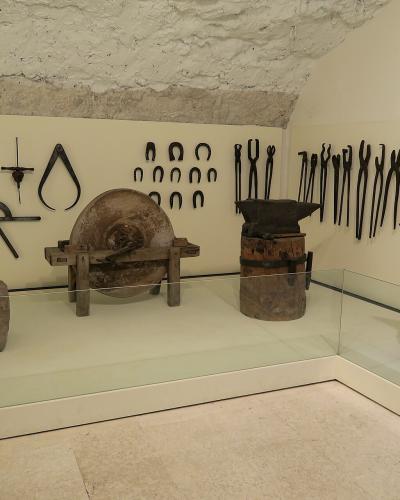 A museum display case containing antique blacksmithing tools, including tongs, horseshoes, an anvil, and a grinding wheel, mounted on a wall beneath a rough stone ceiling.