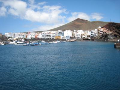 A coastal town with white buildings, a harbor filled with small boats, and a prominent conical volcanic hill rising behind the settlement under a partly cloudy sky.