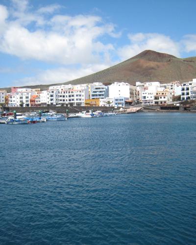 A coastal town with white buildings, a harbor filled with small boats, and a prominent conical volcanic hill rising behind the settlement under a partly cloudy sky.