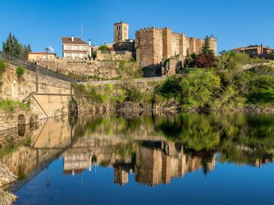 A stone castle and adjacent buildings on a hill overlooking a river with a clear reflection in the water.