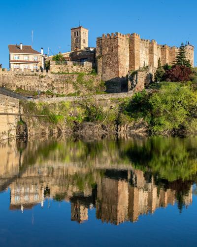 A stone castle and adjacent buildings on a hill overlooking a river with a clear reflection in the water.