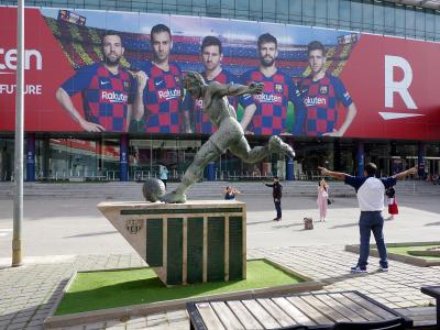A bronze statue of a football player kicking a ball stands on a stone plinth in front of a large building facade covered by a massive red advertising banner featuring FC Barcelona players and the "Rakuten" sponsor logo.