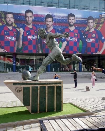 A bronze statue of a football player kicking a ball stands on a stone plinth in front of a large building facade covered by a massive red advertising banner featuring FC Barcelona players and the "Rakuten" sponsor logo.