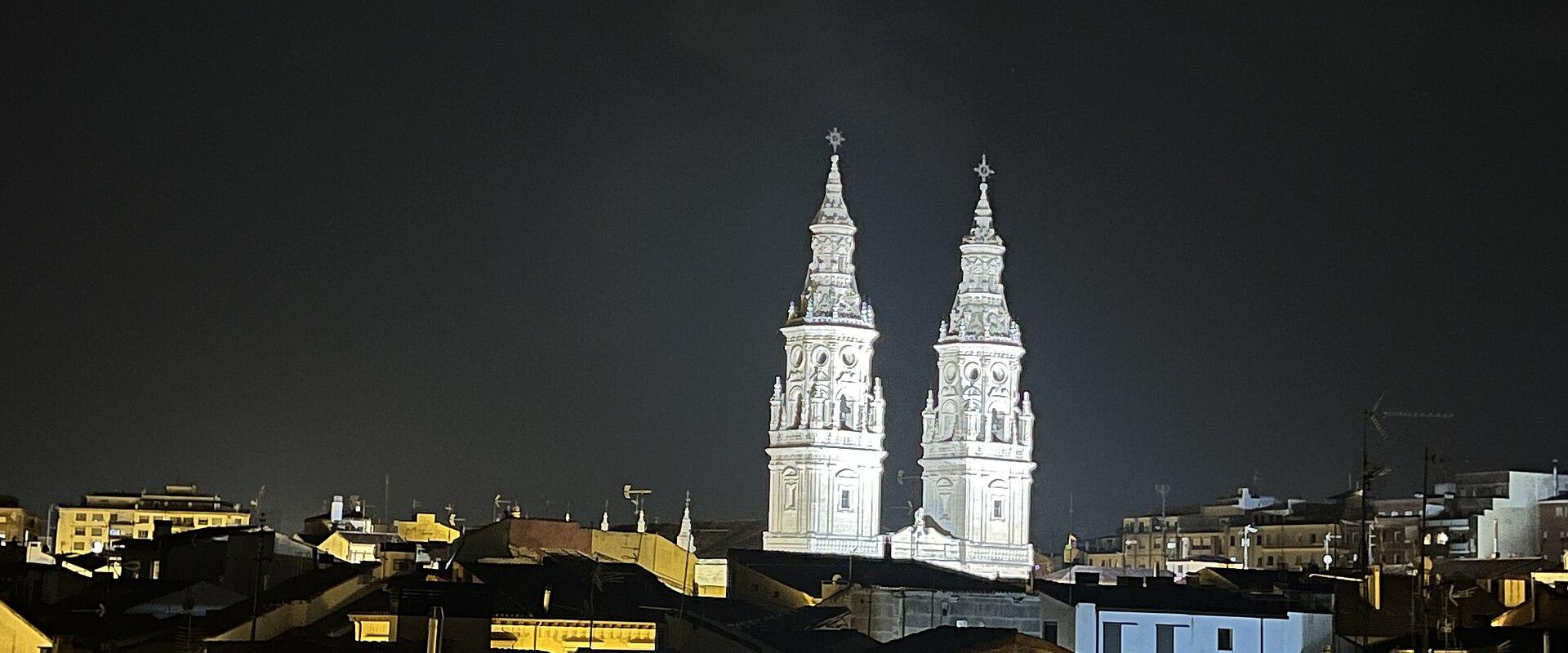 A night photograph showing two illuminated, ornate white bell towers rising above the dark rooftops of a city.