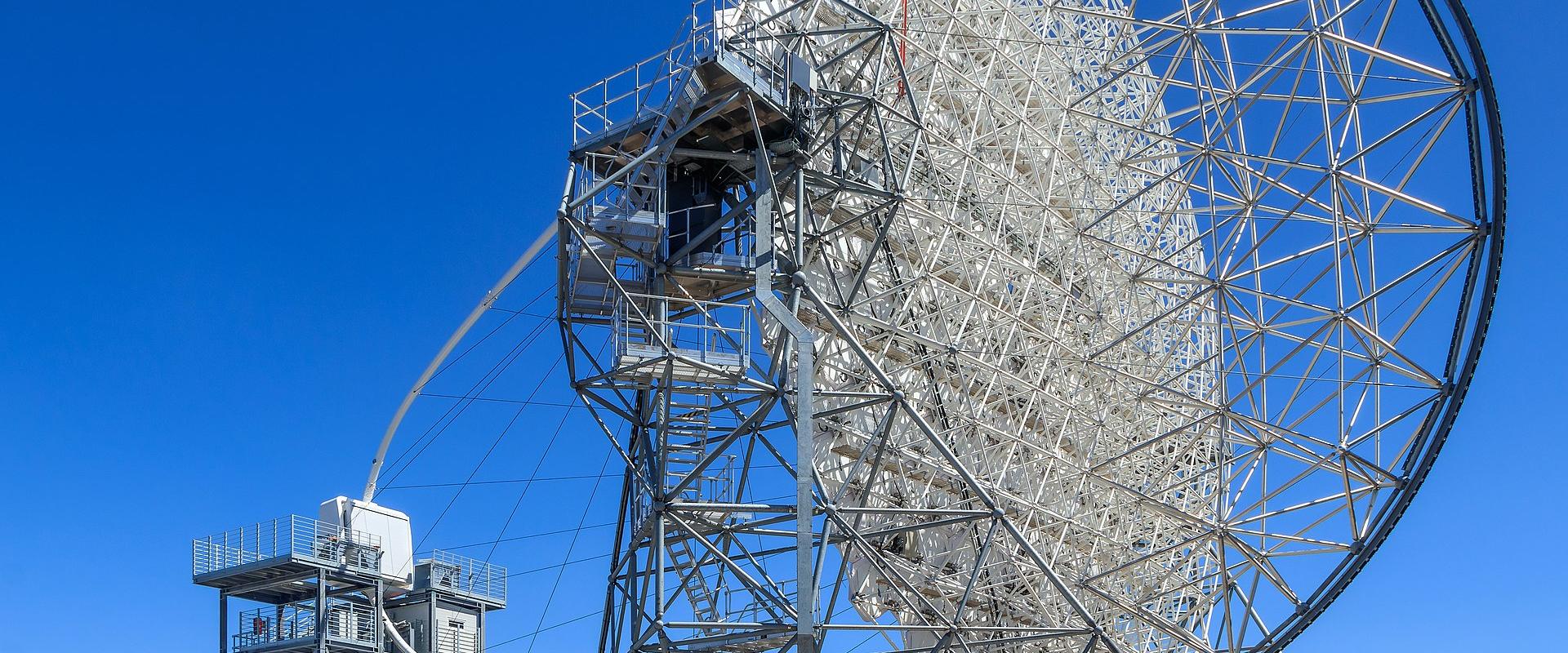 A large white radio telescope dish on a metal support structure against a clear blue sky, with a smaller adjacent tower and fencing in the foreground.