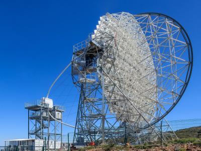 A large white radio telescope dish on a metal support structure against a clear blue sky, with a smaller adjacent tower and fencing in the foreground.