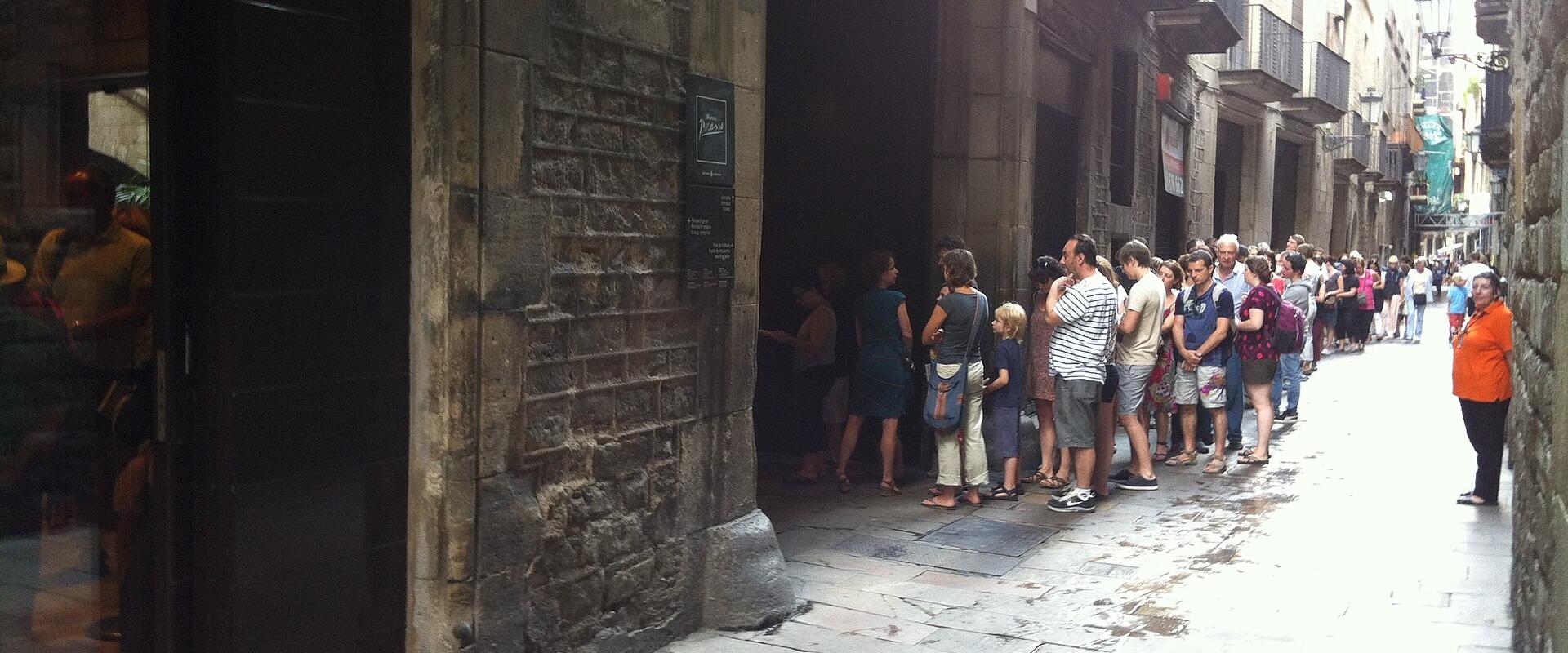 A line of tourists waiting outside the arched entrance of a historic stone building on a narrow cobblestone street, with a black sign mounted on the wall next to the door.