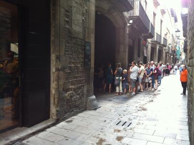 A line of tourists waiting outside the arched entrance of a historic stone building on a narrow cobblestone street, with a black sign mounted on the wall next to the door.