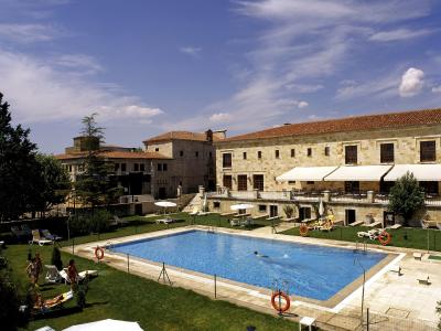A rectangular swimming pool surrounded by a grassy lawn, lounge chairs, and umbrellas, with a large, multi-story stone building featuring a terracotta tile roof and white awnings in the background under a blue sky.
