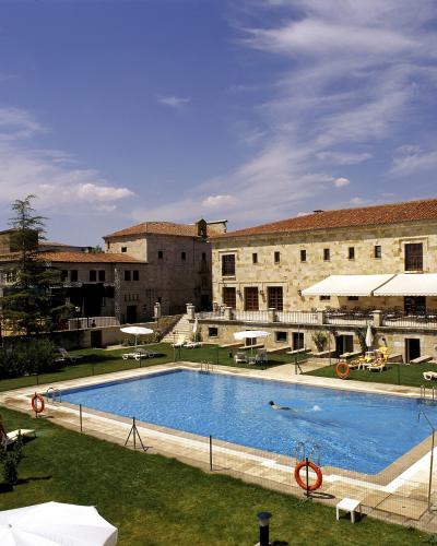 A rectangular swimming pool surrounded by a grassy lawn, lounge chairs, and umbrellas, with a large, multi-story stone building featuring a terracotta tile roof and white awnings in the background under a blue sky.