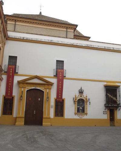 A white and yellow building facade with two red vertical banners reading "2013 AÑO JUBILAR MUSEO" and "2014 AÑO JUBILAR MUSEO", a central arched wooden door, a religious niche with a statue, and a plaque on the right wall that appears to read "PLAZA 