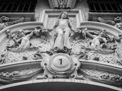 A black and white photograph of an ornate stone relief sculpture above an entrance, featuring a central female figure flanked by cherubs and decorative elements, with the number '1' prominently displayed in a circular medallion at the bottom center.