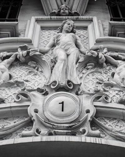 A black and white photograph of an ornate stone relief sculpture above an entrance, featuring a central female figure flanked by cherubs and decorative elements, with the number '1' prominently displayed in a circular medallion at the bottom center.