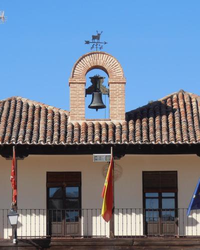 A white municipal building with a terracotta tiled roof, a central brick bell tower, and three flags (Spanish, regional, and EU) hanging from the facade.