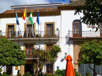 A white, two-story building with a tiled roof, balconies, and four flags (Andalusia, Spain, Marbella, EU) flying from the facade, surrounded by trees and an orange umbrella.