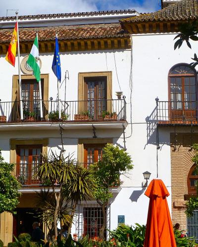 A white, two-story building with a tiled roof, balconies, and four flags (Andalusia, Spain, Marbella, EU) flying from the facade, surrounded by trees and an orange umbrella.