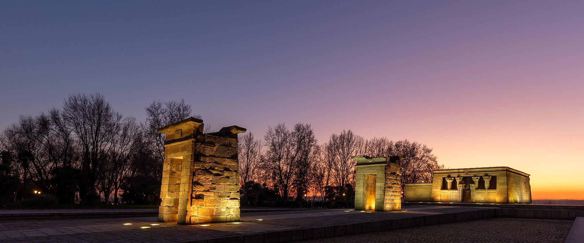 An ancient Egyptian temple complex featuring a pylon gate and a main sanctuary building, illuminated at twilight against a colorful sky.