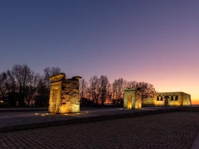An ancient Egyptian temple complex featuring a pylon gate and a main sanctuary building, illuminated at twilight against a colorful sky.