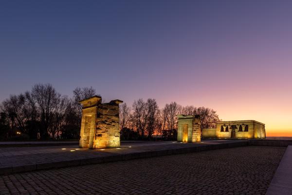 An ancient Egyptian temple complex featuring a pylon gate and a main sanctuary building, illuminated at twilight against a colorful sky.