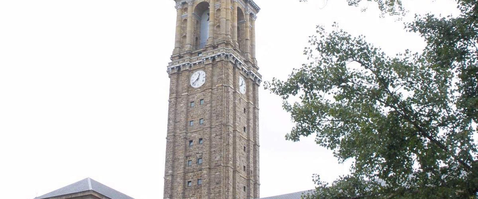 A tall, historic stone clock tower with a spire and a cross, attached to a large, multi-story stone building with rows of rectangular windows, viewed from a low angle with trees on the right and parked cars in the foreground.