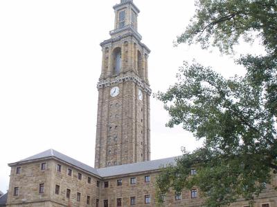 A tall, historic stone clock tower with a spire and a cross, attached to a large, multi-story stone building with rows of rectangular windows, viewed from a low angle with trees on the right and parked cars in the foreground.