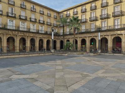 A large, rectangular urban plaza paved with grey stone tiles, surrounded by multi-story buildings with arcades on the ground floor and balconies above, featuring palm trees in the center.