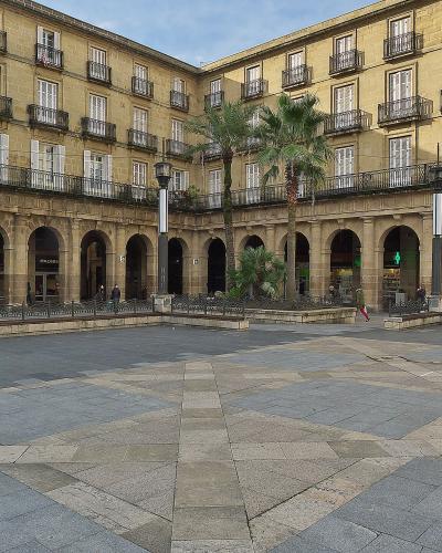A large, rectangular urban plaza paved with grey stone tiles, surrounded by multi-story buildings with arcades on the ground floor and balconies above, featuring palm trees in the center.