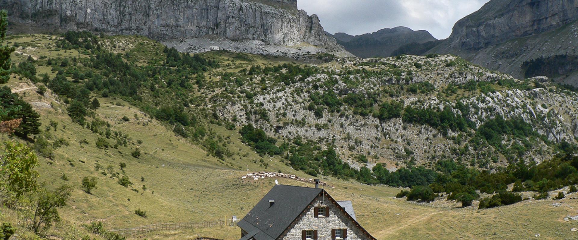 A stone mountain refuge building with a dark roof sits in a grassy valley surrounded by steep limestone cliffs and rocky slopes under a cloudy sky.