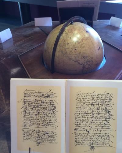 An open historical manuscript with cursive handwriting and a terrestrial globe on a wooden display table inside a museum.
