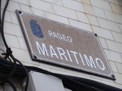 A close-up of a rectangular street sign mounted on a white tiled wall, displaying the text "PASEO MARITIMO" in white capital letters alongside a blue coat of arms featuring a lighthouse.