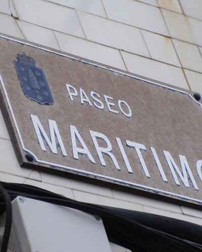 A close-up of a rectangular street sign mounted on a white tiled wall, displaying the text "PASEO MARITIMO" in white capital letters alongside a blue coat of arms featuring a lighthouse.