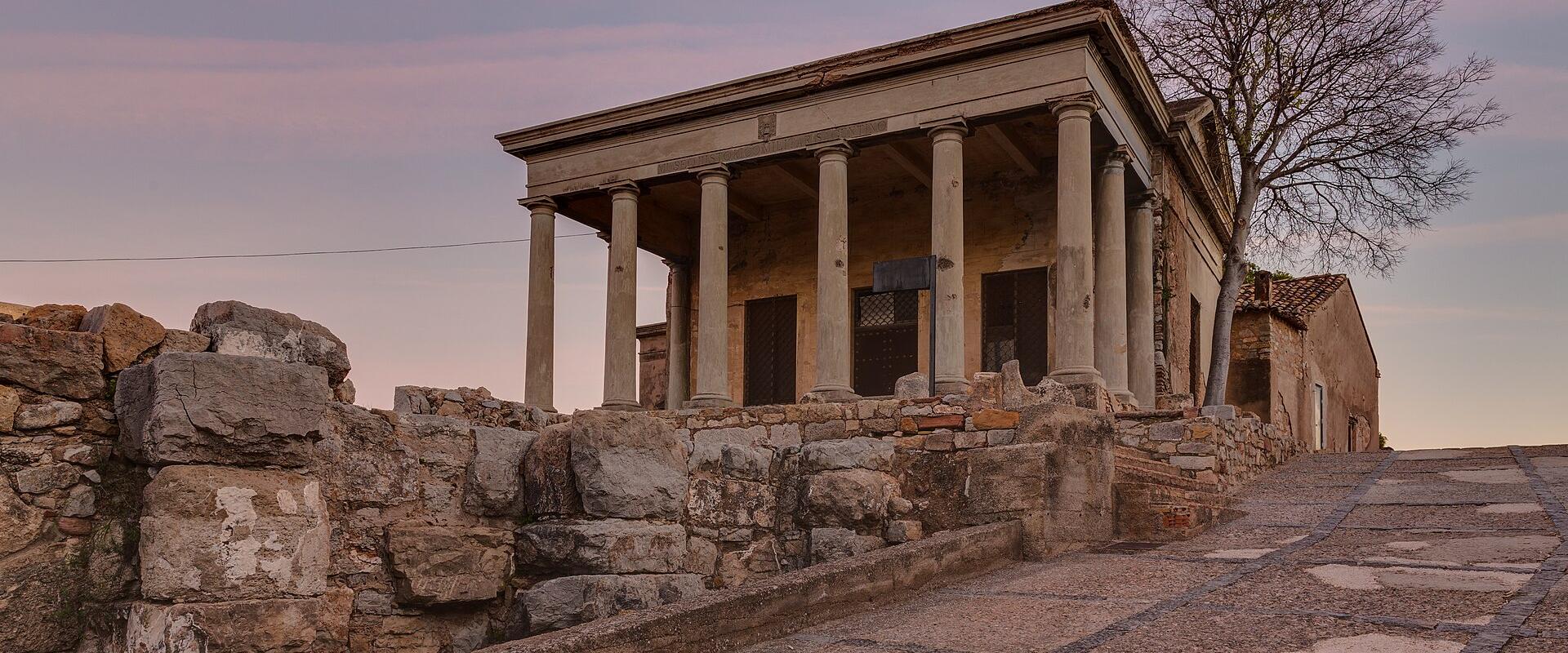 A neoclassical temple-like structure with a portico of columns, situated atop ancient stone ruins and a paved ramp, set against a twilight sky.