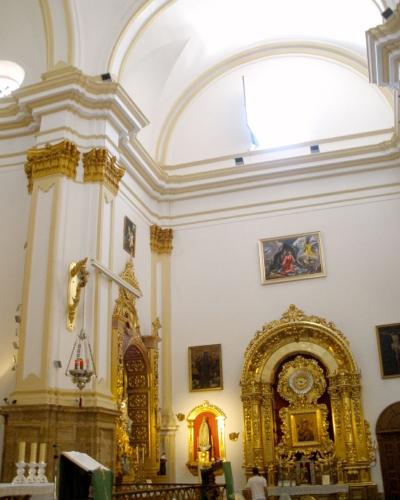 The interior of a church featuring white walls, ornate gold altarpieces, religious paintings, and a high arched ceiling with a flag hanging near the dome.