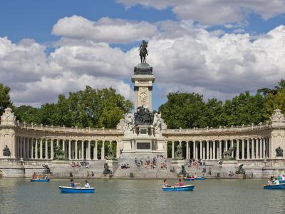 The image shows the Monument to Alfonso XII located on the island in the center of the Estanque Grande (Great Pond) within a large public park, with people rowing boats in the foreground.