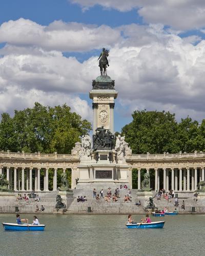 The image shows the Monument to Alfonso XII located on the island in the center of the Estanque Grande (Great Pond) within a large public park, with people rowing boats in the foreground.