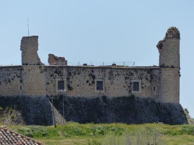 A stone ruin with two prominent, damaged towers and a long wall featuring three rectangular windows, set against a clear blue sky with green vegetation in the foreground.