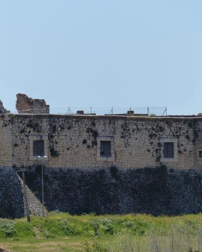 A stone ruin with two prominent, damaged towers and a long wall featuring three rectangular windows, set against a clear blue sky with green vegetation in the foreground.