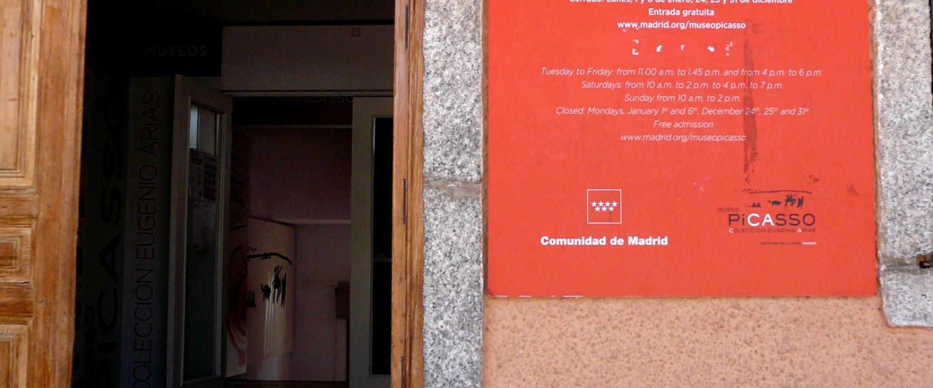 A close-up view of the entrance to a museum, featuring a large red sign on the wall that reads "PICASSO MUSEO COLECCIÓN EUGENIO ARIAS" along with opening hours and the logo for the Madrid regional government.