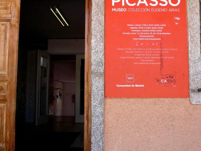 A close-up view of the entrance to a museum, featuring a large red sign on the wall that reads "PICASSO MUSEO COLECCIÓN EUGENIO ARIAS" along with opening hours and the logo for the Madrid regional government.