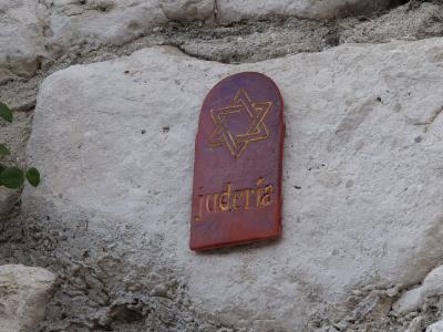 A close-up of a reddish-brown, tombstone-shaped plaque mounted on a rough, light-colored stone wall, featuring a gold Star of David and the word "judería" in gold lettering.