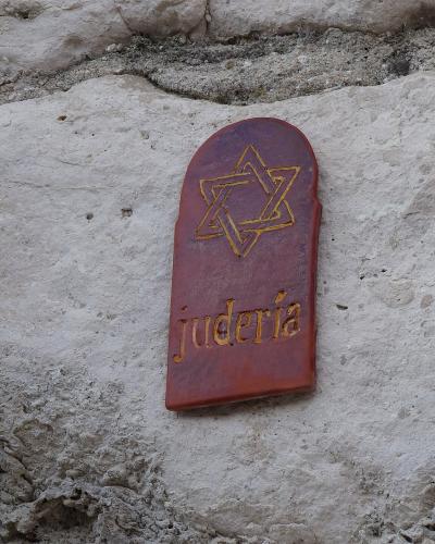 A close-up of a reddish-brown, tombstone-shaped plaque mounted on a rough, light-colored stone wall, featuring a gold Star of David and the word "judería" in gold lettering.