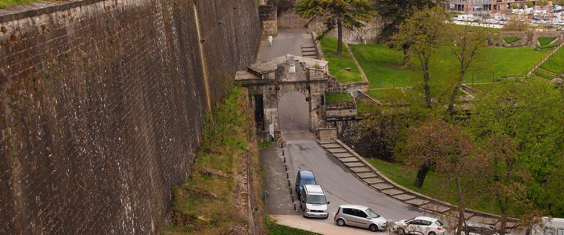 A high-angle view of a massive stone fortification wall with a grassy rampart, featuring a prominent arched gateway (Puerta de Santa Engracia) and a road with cars passing through it.