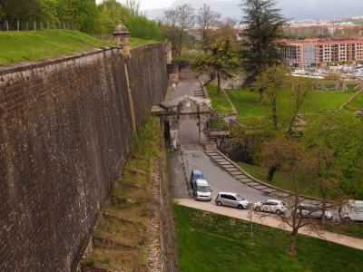 A high-angle view of a massive stone fortification wall with a grassy rampart, featuring a prominent arched gateway (Puerta de Santa Engracia) and a road with cars passing through it.