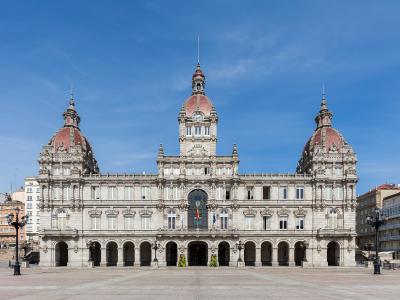 A large, ornate neoclassical building with three prominent domes and a central clock tower, situated in a wide paved plaza under a clear blue sky.