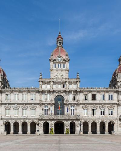 A large, ornate neoclassical building with three prominent domes and a central clock tower, situated in a wide paved plaza under a clear blue sky.