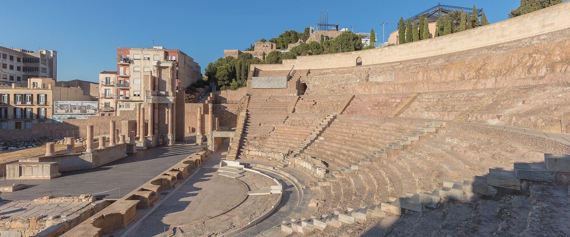 A wide-angle view of the excavated Roman theatre ruins featuring tiered stone seating, a stage area with reconstructed columns, and modern apartment buildings in the background.