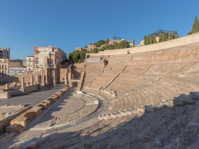 A wide-angle view of the excavated Roman theatre ruins featuring tiered stone seating, a stage area with reconstructed columns, and modern apartment buildings in the background.